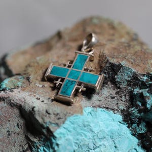 Może przedstawiać: Srebrny wisiorek w kształcie krzyża z turkusową inkrustacją. Krzyż ma tradycyjny wzór z centralnym kwadratem i czterema ramionami.