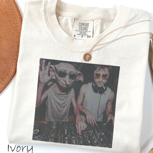 May include: Ivory-colored Comfort Colors t-shirt featuring a graphic of two figures wearing sunglasses and operating DJ equipment. A gold necklace with a pendant rests on the shirt. The shirt is labeled "S P CH".