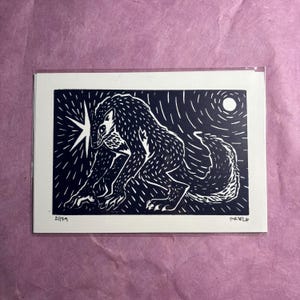 Op de afbeelding: Een zwart-witte linosnede op papier met een gestileerd wezen dat lijkt op een weerwolf in een gehurkte houding. Het kunstwerk bevat een heldere ster aan de linkerkant, een cirkelvormige maan aan de rechterkant en de handgeschreven tekst 21/29 en FR026.