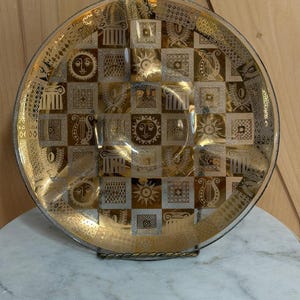 May include: A round glass plate featuring a gold and silver checkerboard pattern. Each square contains detailed illustrations of sun faces, lyres, columns, and laurel wreaths. The plate has a reflective metallic finish and a clear glass border.