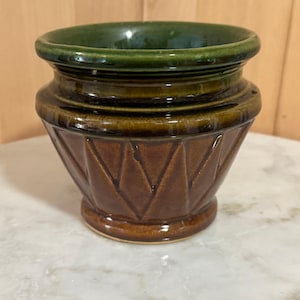 Puede incluir: Pequeño jarrón decorativo de cerámica, marrón por fuera y verde por dentro. El jarrón tiene un patrón geométrico y una base redondeada. El borde es de color verde oscuro.