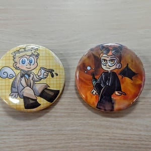May include: Two round button pins with cartoon characters. The first pin features a blonde character with a halo and wings, wearing a brown coat and holding a cup of coffee. The background is a yellow and white checkered pattern. The second pin features a character with red hair and horns, wearing a black coat and holding a bottle. The background is a swirling orange and black pattern.
