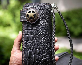 Pode incluir: Uma carteira de couro preta com um padrão de crocodilo e um detalhe metálico em forma de estrela. A carteira tem uma alça de couro trançada.