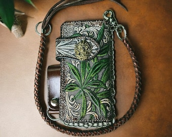 Pode incluir: Uma carteira de couro marrom com um design de folha de cannabis verde, uma fivela decorativa e uma alça trançada. A carteira tem uma corrente prateada e um pequeno acessório de couro marrom. O texto "Leathergood Studio" é visível.
