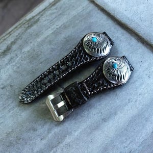 May include: Two black leather watch bands with silver eagle head buckles and turquoise accents. The bands have a woven pattern.