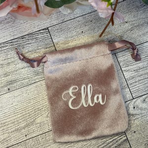 May include: A dusty rose velvet pouch with the name "Ella" embroidered in white script. The drawstring bag is tied with a matching satin ribbon. Pink and white flowers are in the background.