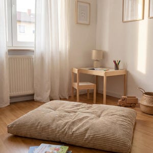 Op de afbeelding: Een rechthoekig vloerkussen van beige corduroy met getufte details, geplaatst op een houten vloer. Een kleine stapel kinderboeken met kleurrijke ge&iuml;llustreerde covers ligt op de vloer bij de rand van het kussen.