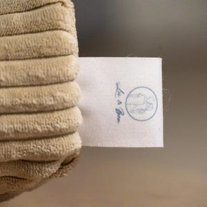 Op de afbeelding: Een beige corduroy stof met een getextureerd, geribbeld oppervlak en een wit rechthoekig kledinglabel. Het label toont een cirkelvormig logo met een lijntekening van een persoon en de tekst Lu & Bee gedrukt in een donkerblauwe inktkleur.