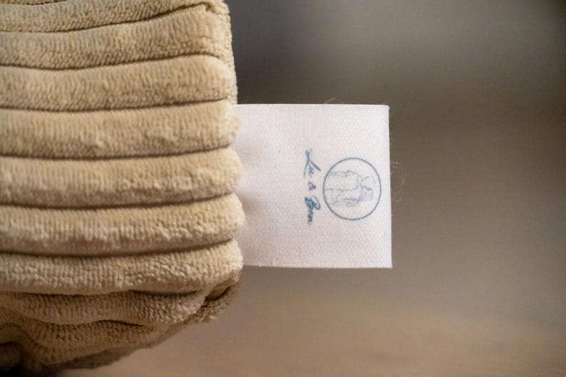 Op de afbeelding: Een beige corduroy stof met een getextureerd, geribbeld oppervlak en een wit rechthoekig kledinglabel. Het label toont een cirkelvormig logo met een lijntekening van een persoon en de tekst Lu & Bee gedrukt in een donkerblauwe inktkleur.