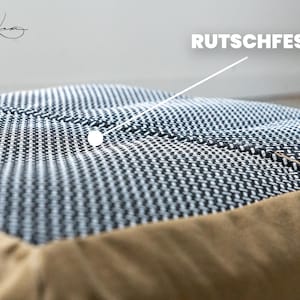 Op de afbeelding: Een vierkant huisdierkussen met een zwart-wit geweven bovenkant met een textuurpatroon. Het kussen heeft een effen beige rand langs de zijkanten. Een witte lijn wijst naar het midden van het kussen met de tekst RUTSCHFEST in het wit.