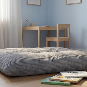 Op de afbeelding: Een groot, rechthoekig vloerkussen bekleed met grijze corduroy stof met een getuft oppervlak. Het kussen heeft een zachte, geribbelde textuur en ligt op een lichte houten vloer, wat zorgt voor een lage zitgelegenheid in de kamer.