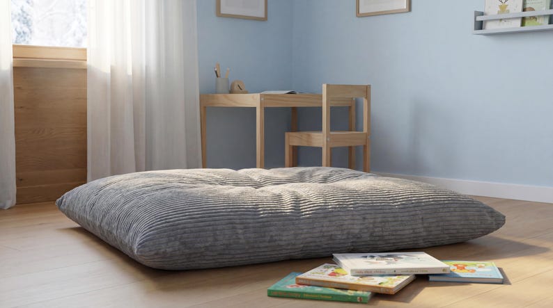 Op de afbeelding: Een groot, rechthoekig vloerkussen bekleed met grijze corduroy stof met een getuft oppervlak. Het kussen heeft een zachte, geribbelde textuur en ligt op een lichte houten vloer, wat zorgt voor een lage zitgelegenheid in de kamer.