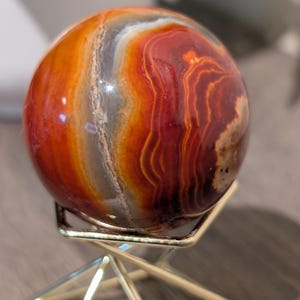 Può includere: Una sfera di agata lucidata, con fasce rosse, arancioni, bianche e grigie. La gemma è esposta su un supporto geometrico in metallo dorato, che mette in risalto i suoi motivi e colori naturali. La sfera di agata è un oggetto decorativo.