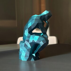 Può includere: Una scultura in 3D stampata in blu turchese di una persona in una posa di pensiero. La scultura è realizzata con molti piccoli triangoli e ha un design geometrico.