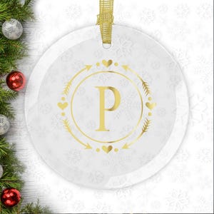 May include: A white round glass ornament with a gold snowflake pattern and a gold circle with the letter "P" in the center. The circle is surrounded by gold arrows and hearts.