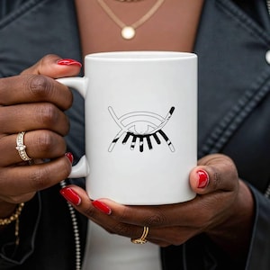 Könnte beinhalten: Weißer Keramikbecher mit einem schwarz-weißen grafischen Augendesign. Das Augendesign hat eine minimalistische Ästhetik. Der Becher wird von einer Person mit rotem Nagellack und Goldschmuck gehalten.