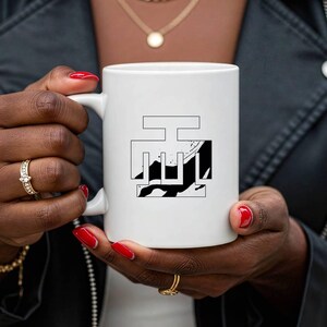 May include: White ceramic mug featuring a black and white graphic design. The design is a stylized, abstract image. The mug is held by a person with red nail polish and gold jewelry.