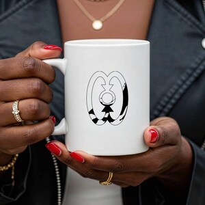May include: White ceramic mug featuring a black and white graphic design. The design includes an abstract figure with a geometric pattern. The mug has a standard handle and is held by a person with red nail polish.