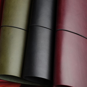 Puede incluir: Cuatro cuadernos rectangulares de cuero dispuestos uno al lado del otro, cada uno asegurado con una banda elástica delgada. Las cubiertas son de colores sólidos que incluyen rojo brillante, verde oliva, negro mate y burdeos oscuro.