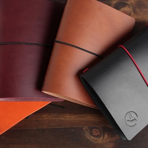 May include: Four leather journals in orange, burgundy, tan, and black. The orange, burgundy, and tan journals are secured with black elastic bands. The black journal features a red elastic band and a circular embossed bird logo on the cover.