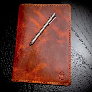 May include: A brown leather journal with a pen resting on top. The journal has a distressed texture and a logo in the bottom right corner. The pen is silver and has a sleek design.