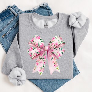 May include: A gray sweatshirt with a pink floral bow design. The bow is made of pink fabric with a floral pattern and is tied in a knot in the center. The sweatshirt is a comfortable and stylish piece of clothing.