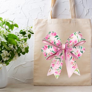 May include: A beige tote bag with a large pink bow printed on the front. The bow is decorated with a floral pattern of pink flowers and green leaves.