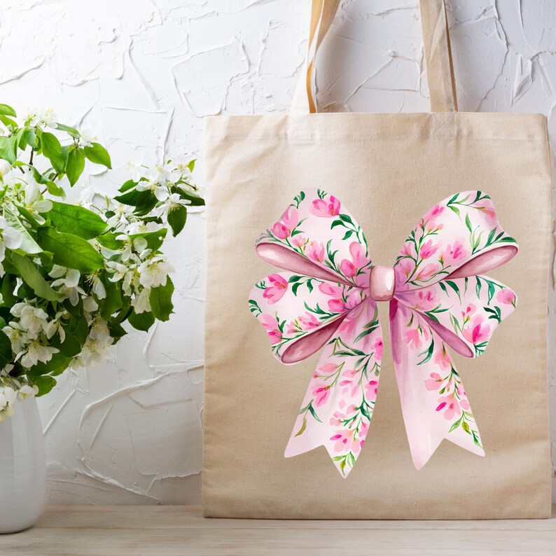 May include: A beige tote bag with a large pink bow printed on the front. The bow is decorated with a floral pattern of pink flowers and green leaves.