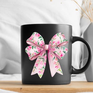 May include: Black coffee mug with a pink floral bow design. The bow is made of pink ribbon with a floral pattern and is tied in a knot.