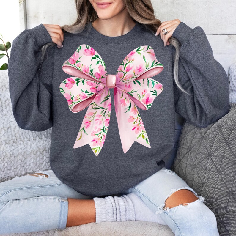 May include: A dark gray sweatshirt with a large pink floral bow design. The bow is made of pink ribbon with a floral pattern and has a large pink gem in the center.