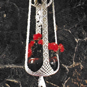 Peut inclure: Un porte-plante en macramé blanc avec un pompon, intitulé "Angel Flake" du livre "Macrame Portraits, Book 2, $1.50" par Vickie.