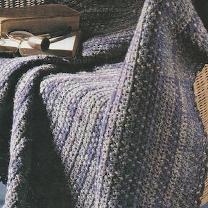 Könnte beinhalten: Eine gestrickte, violett-graue Wolldecke liegt über einem Korbstuhl. Auf dem Sitz des Stuhls liegen ein Buch und eine Lesebrille.