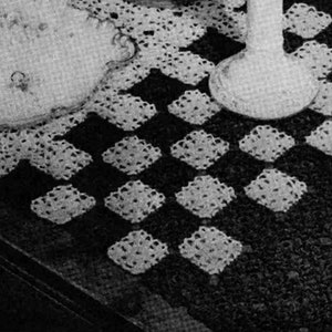 Op de afbeelding: Een zwarte tafel met een wit gehaakt tafellaken. Twee witte kandelaars met kaarsen staan op de tafel. Een kleine, rechthoekige doos staat op de tafel.