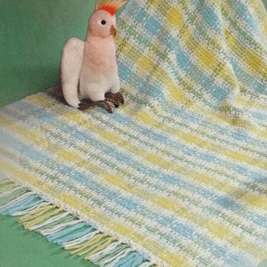 Puede incluir: Una manta de bebé de ganchillo con un patrón a rayas en tonos de azul, amarillo y blanco. La manta tiene un borde de flecos y se muestra sobre una superficie verde con un pájaro de peluche rosa y blanco.