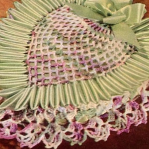 Könnte beinhalten: Eine gehäkelte herzförmige Tischdecke mit einer grünen Schleife aus Satinband. Die Tischdecke ist aus weißem und violettem Garn gefertigt und hat ein zartes Spitzenmuster.