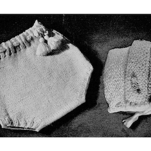 Op de afbeelding: Een gebreide baby muts en luier set in wit. De muts heeft een eenvoudig ontwerp en de luier heeft een gerimpelde rand met kwastjes.