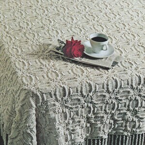 May include: Cream-colored crocheted tablecloth with a detailed pattern and fringe. A red rose, a cup of coffee, and a newspaper are placed on the tablecloth, creating a cozy and inviting scene. The intricate crochet work adds texture and visual interest.