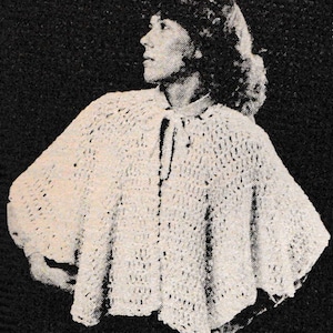 May include: A cream-colored, crocheted cape with a tie closure at the neck. The cape has a textured, open weave design and a wide, flowing shape. The background is a solid black.