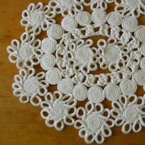 Op de afbeelding: Een witte, cirkelvormige doily met een bloemenontwerp. De doily heeft een herhalend patroon van spiraal- en bloembladachtige elementen, waardoor een open textuur ontstaat. Het handgemaakte stuk is op een houten oppervlak geplaatst.