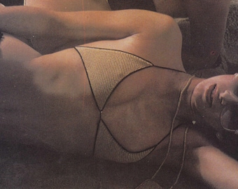 May include: A woman wearing a gold crochet bikini and sunglasses is lying down. The bikini has a geometric design with black trim. The woman is on a sandy surface, possibly a beach. The image has a vintage aesthetic.