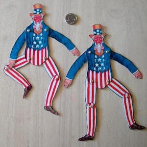 May include: Two cardboard Uncle Sam figures with red, white, and blue striped pants and jackets. The figures are posed with their legs crossed and their arms outstretched.