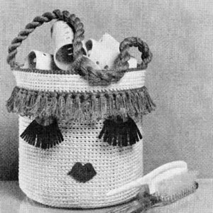 May include: A white crocheted basket with a rope handle and a face design. The face has black eyelashes, a red lip, and brown tassels around the top edge. A white comb is lying next to the basket.