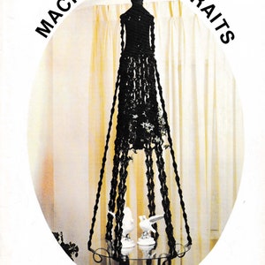 以下が含まれることがあります： 黒いマクラメのプラントハンガー。ガラスの棚には白い鳥の置物が2つ置かれています。「Macrame Portraits」というタイトルの本からの画像で、Sir Lancelotによるものです。プラントハンガーはクリーム色のカーテンの前に吊り下げられています。