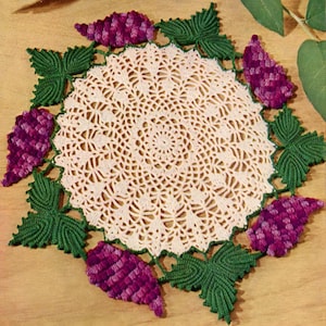 May include: A crocheted doily with a delicate floral design featuring pink and white yarn. The doily is surrounded by a border of green leaves and purple grapes.