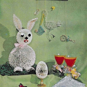 May include: A white crocheted bunny, a crocheted bird mobile, and two glasses of red liquid on a green tablecloth. The bunny has pink ribbon around its neck and a crocheted egg sits in front of it.