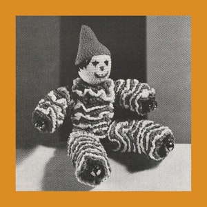 May include: A black and white crocheted clown doll with a pointed hat and bells on its feet. The doll is sitting on a white surface.