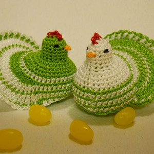 May include: Two crocheted green and white chickens with red combs. The chickens are sitting on a white surface with yellow jelly beans.