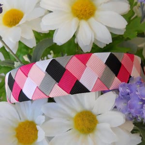 May include: A woven headband with pink, white, black, and gray ribbon. The headband is braided in a diagonal pattern.
