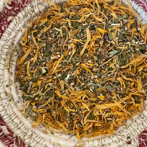 May include: A close-up of a bowl filled with a blend of dried herbs and flowers, including yellow calendula petals, green leafy herbs, and other dried botanicals.