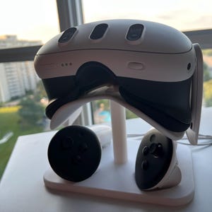 May include: A white and black VR headset with three buttons on top, resting on a white stand. Two black controllers with buttons are positioned below. The setup is on a white surface near a window.